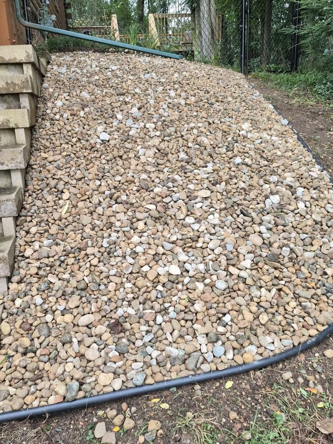 Rounded gravel bed with landscape edging and a green corrugated drainage pipe beside a concrete block retaining wall.