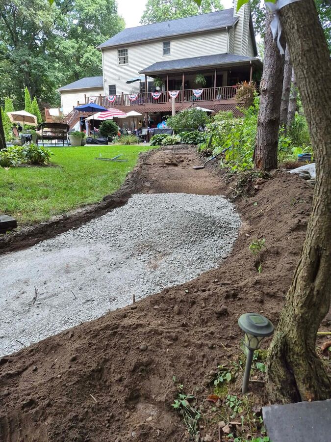 A crushed stone base laid along an excavated route through a backyard garden beside a raised deck.
